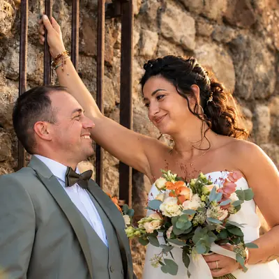 Les mariés complices devant un mur de pierre, bouquet à la main